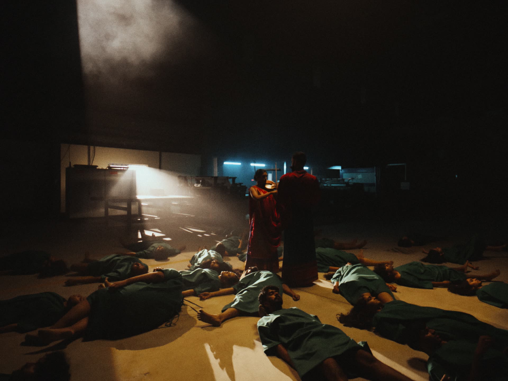 Two musicians stand under a single beam of light among figures lying on a factory floor