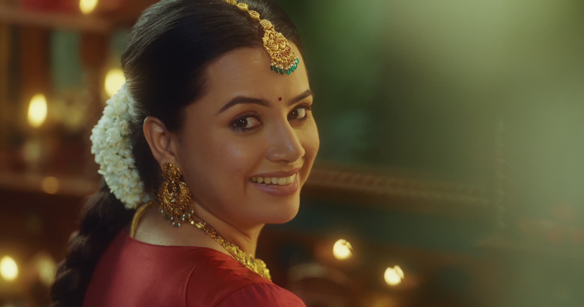 A bride in red silk and gold jewellery smiles over her shoulder, diyas glowing behind her